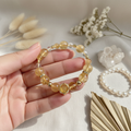 Hand holding a beaded bracelet with a decorative clasp on a neutral background with flowers and leaves.